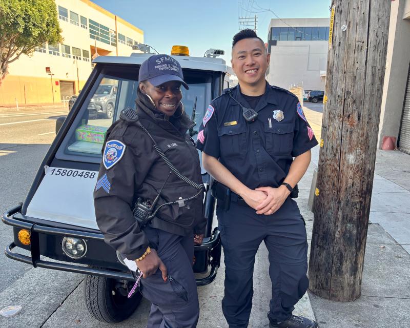 Two people in parking control uniforms stand in front of a city vehicle.