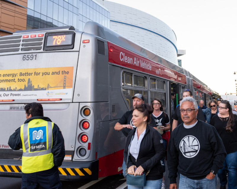 Several Muni riders exit a bus an SFMTA staffer in a yellow vest stands nearby.