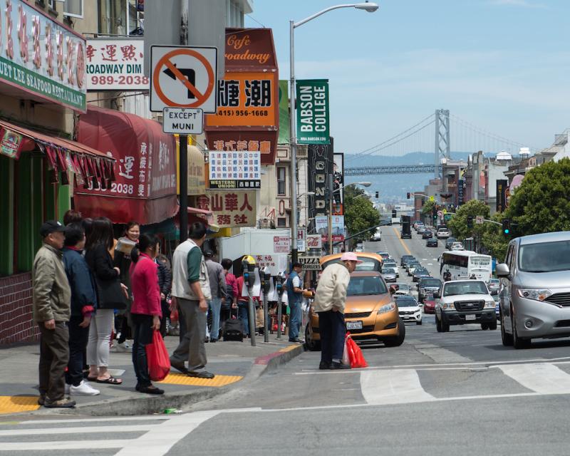 Photo of an intersection with a bridge in the background. Several pedestrians stand on the corner, waiting to cross the crosswalk.