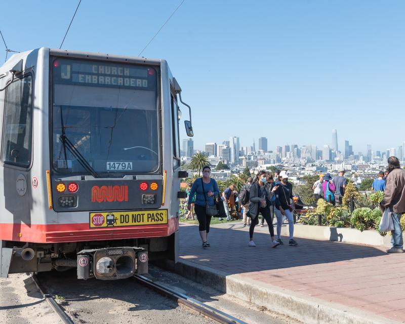 People near a train and light rail tracks with the skyline in the background.