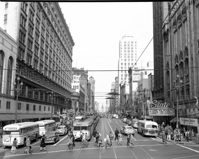 Black and white photo of a busy street in San Francisco. People cross the street as buses and trains wait in traffic.