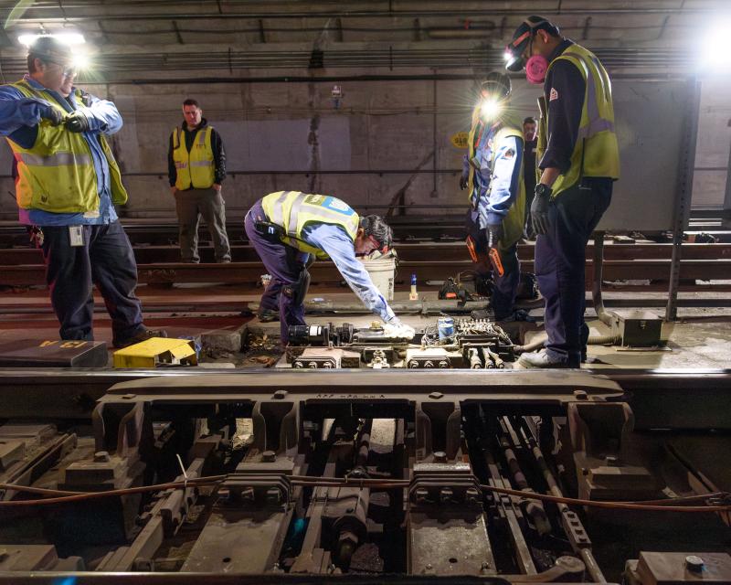 SFMTA crews wearing yellow vests complete work on rails in a Muni Metro tunnel.