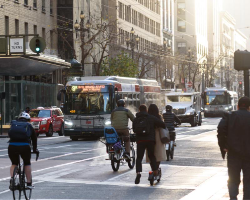 People bike and walk on Market Street as Muni buses near an intersection.