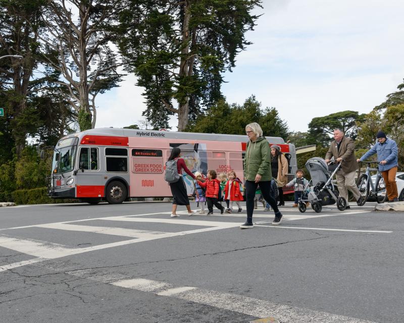 A bus crosses a street as several children and adults pushing strollers walk in an intersection.
