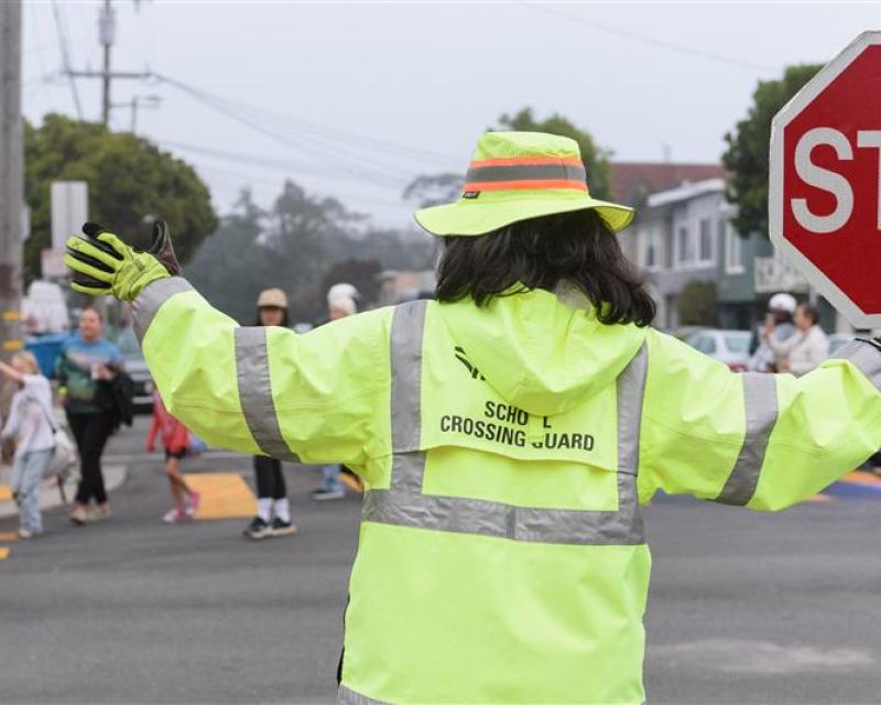 Crossing guard wearing a bright yellow jacket helps people cross a street.