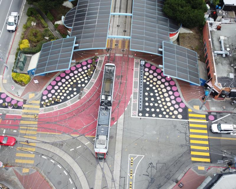 Aerial view of a train entering the West Portal Station Tunnel , yellow crosswalks, new street mural of polka dots, and red painted muni lane all visible