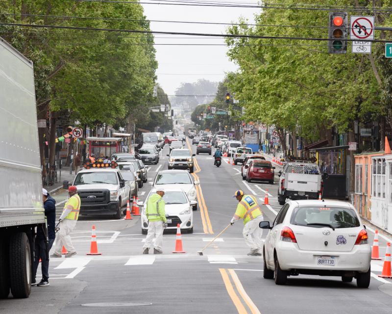 Paint crew workers installing a crosswalk on Valencia street , cars and other pedestrians visible. Parklets and a side running bike lane shown in the distance. 