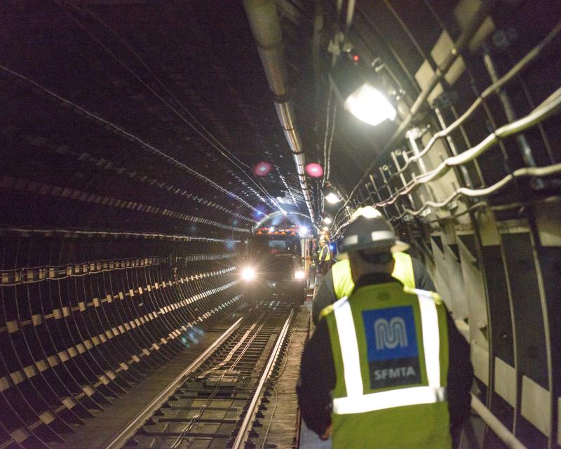 Two people wearing yellow safety vests walk in a Muni tunnel toward a vehicle on the tracks.