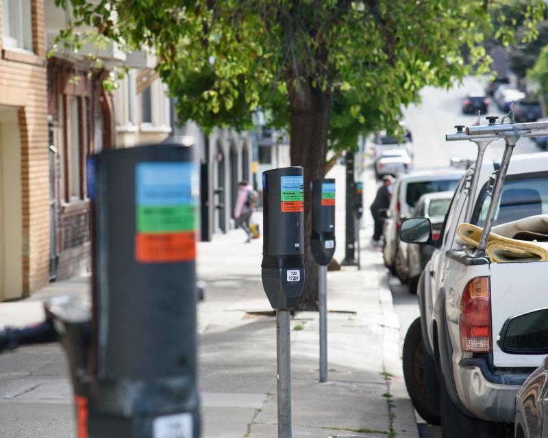 Closeup of SFMTA parking meters on a sidewalk near parked cars.