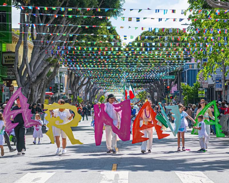  People marching in the street in a parade, holding up letters that spell Carnaval. 
