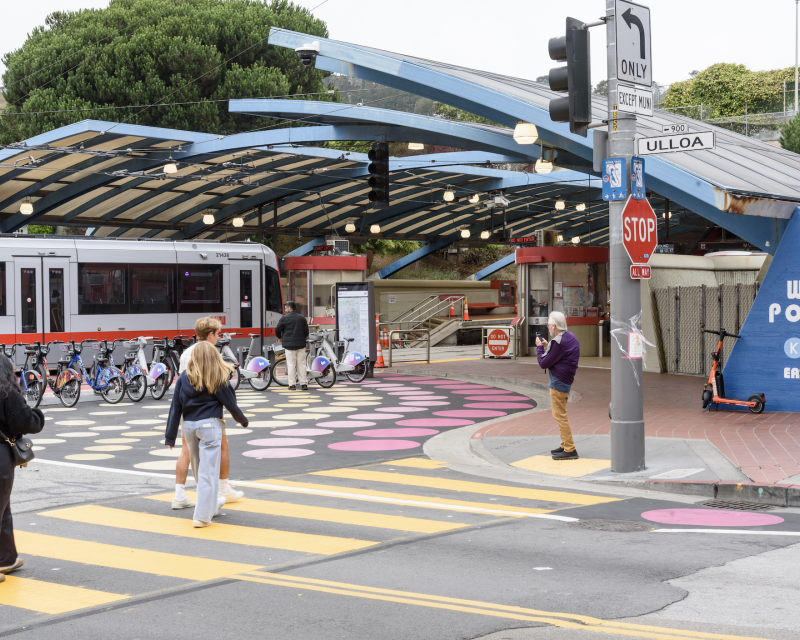 Outside of West Portal Station, several people cross the street near a street mural and bikeshare station while a train pulls in.