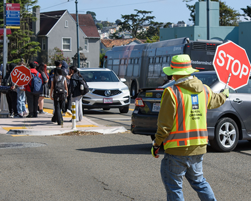 SFMTA Crossing Guard assisting pedestrian safety at intersection with cars and a Muni Metro Stop
