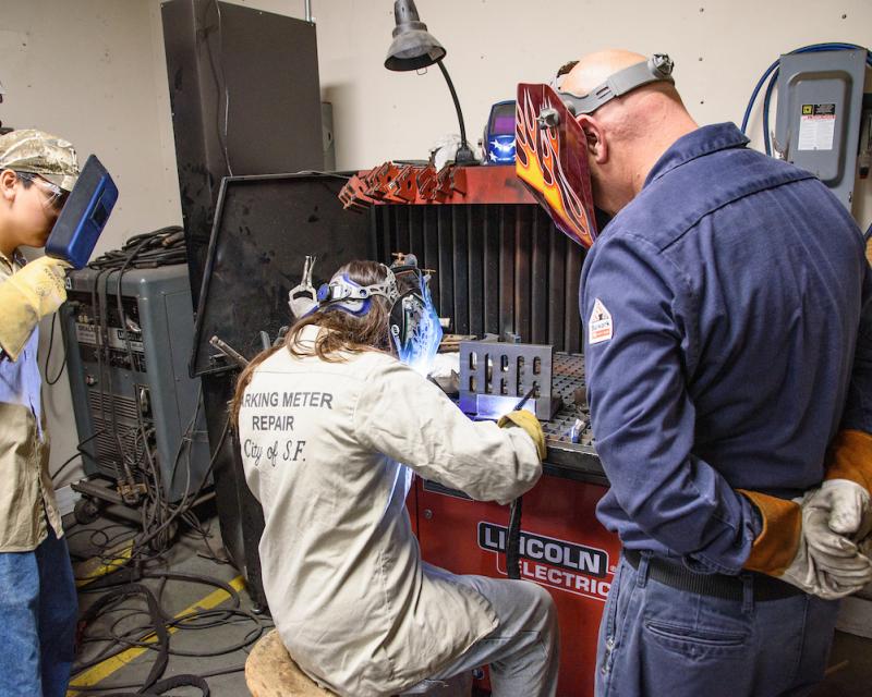 People in work uniforms wearing welders’ masks working on a piece of equipment.