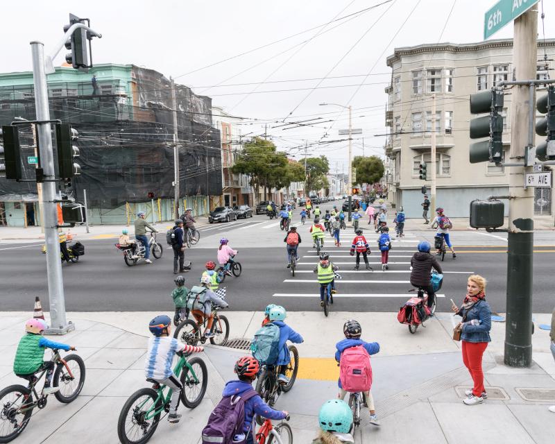Students wearing helmets and safety vests bike across a street during a school event.