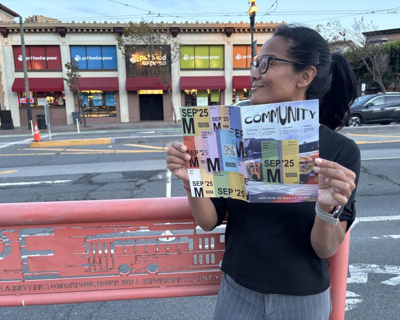 A woman standing near a street and holding a magazine. A colorful building is behind her. 