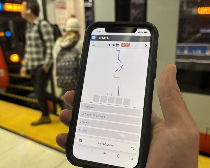 A hand holding a mobile phone in a metro station. A train is in the background.