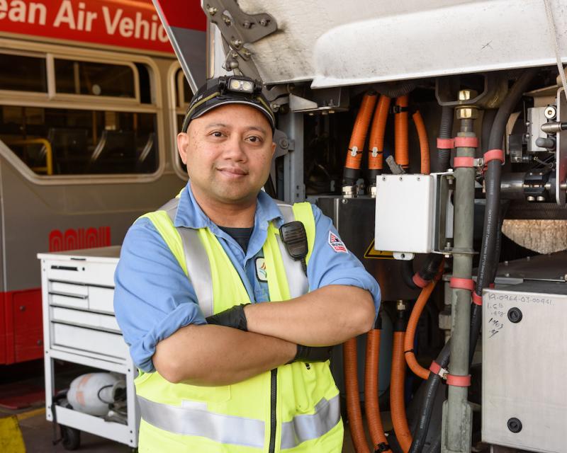 A person wearing a yellow vest and hat while standing near equipment and a transit vehicle.  