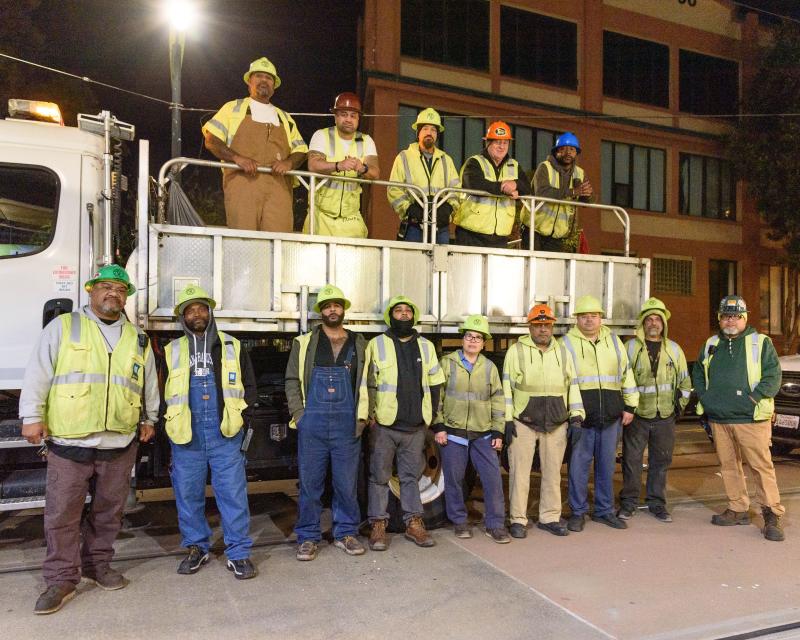 members of Muni's Track Maintenance Team wearing hard hats and standing in a street in front of a truck at night..