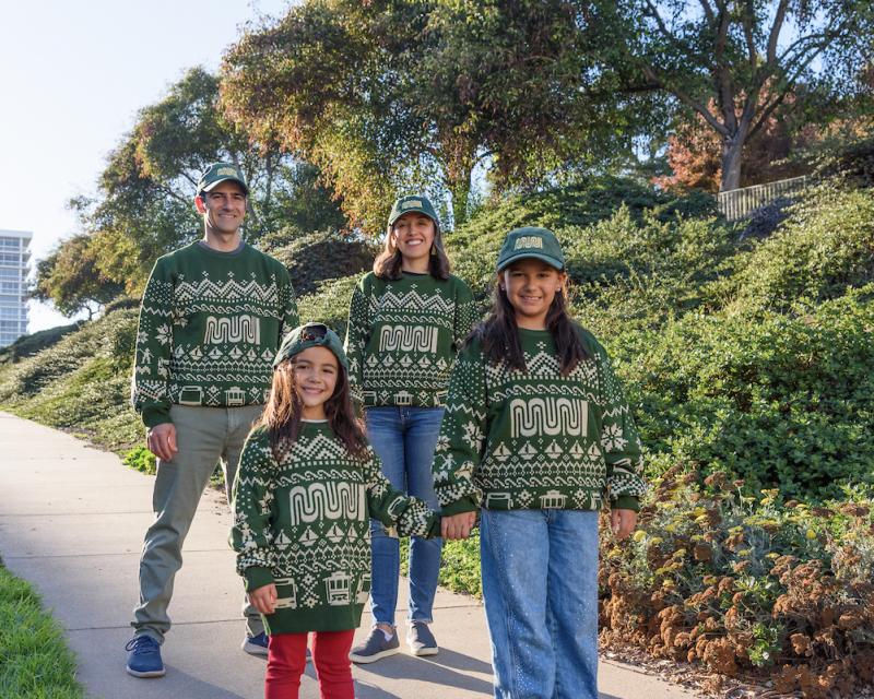 Two adults and two children standing in a park while wearing decorative sweaters and green baseball caps. 
