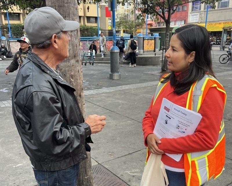 Two people talking on a wide sidewalk. One is wearing an orange vest and sweatshirt. The other is wearing a black jacket and baseball cap.