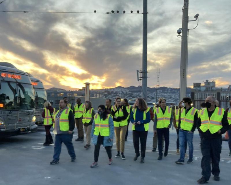 A group of people are pictured walking near buses with a sunset-filled sky in the background.