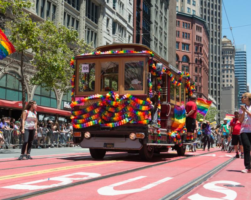 Color photograph from June 26, 2016 showing Muni motorized cable car 62 decorated in rainbow banners on Market Street as part of Pride parade.