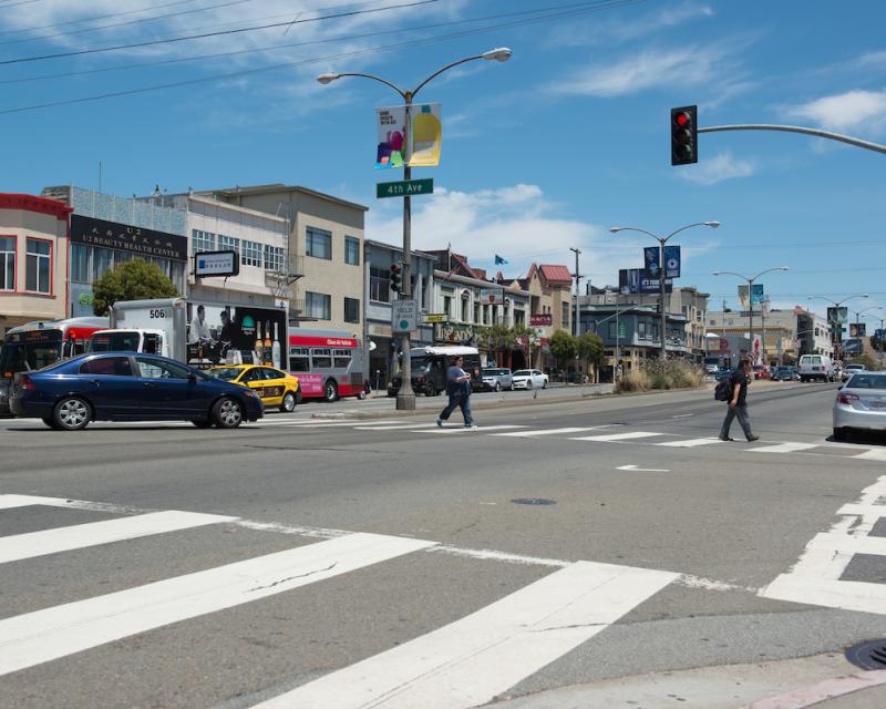 Photograph of Geary Boulevard at 4th Avenue where two pedestrians are crossing in the crosswalk while a car makes a left turn.