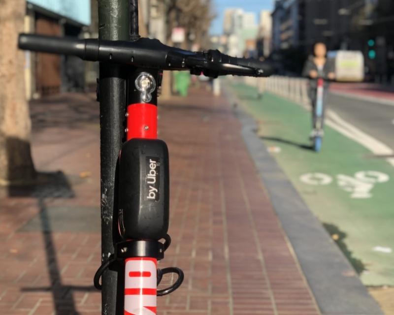 A scooter chained up and a scooter being ridden on the bike path.