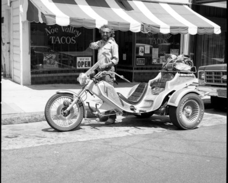 Vintage black and white photo of a person standing over a motorcycle in front of a restaurant "Noe Valley Tacos"