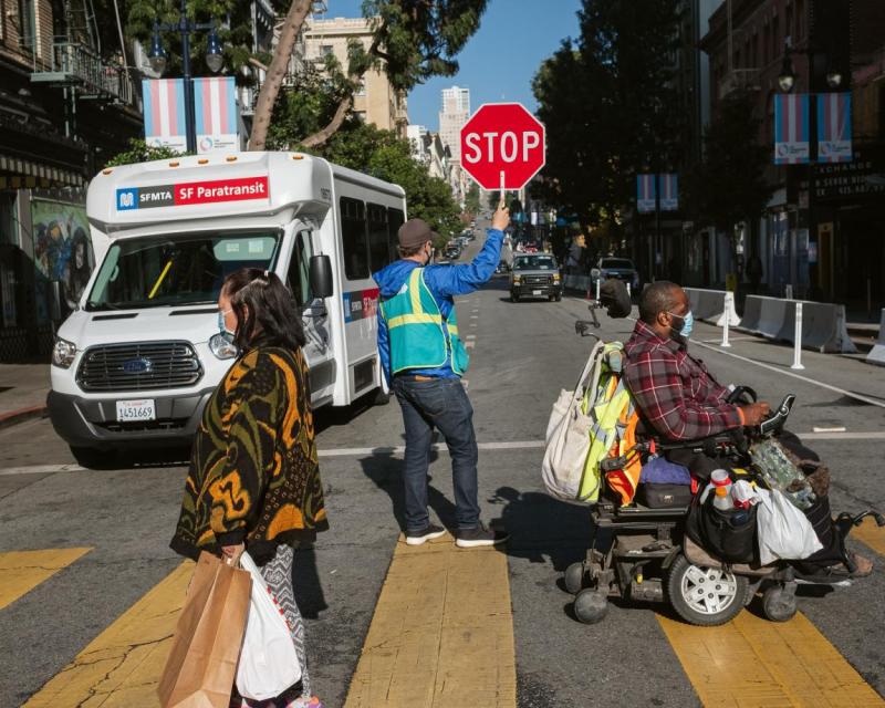 person in wheel chair and person walking, crossing street with crossing gaurd with Paratransit bus in background