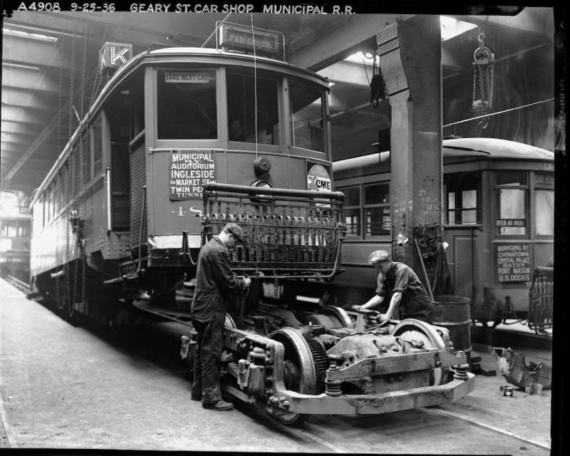 Inside the shops at Geary Car House, two mechanics work on a truck (the combined wheels, suspension, and motor of the streetcar)