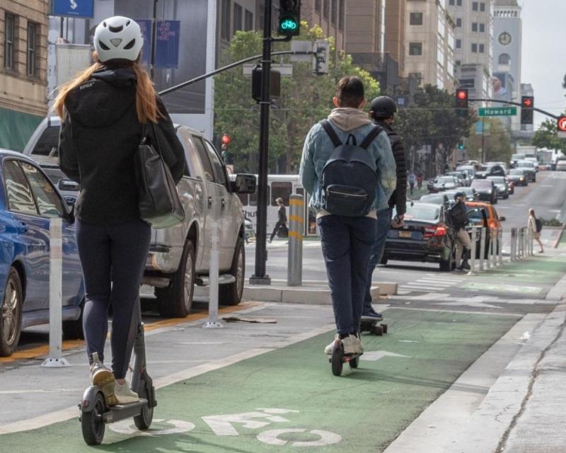 photo of 2 individuals riding powered scooters, one riding a skateboard, and pedestrians