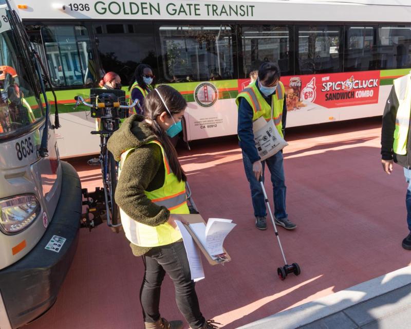 Two staff are shown taking measurements next to a Muni bus on the transit lane. One holds a rolling measuring device. The other 