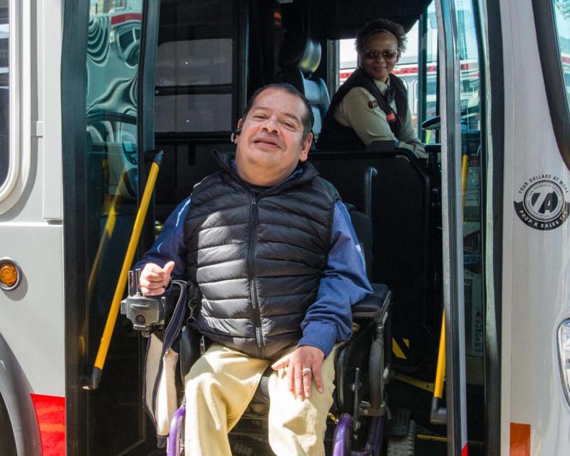 A customer in a wheelchair exits a bus while a driver deploys the lift