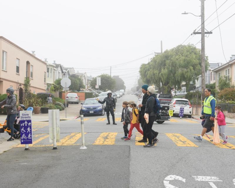 Children, parents and a bicyclist seen crossing in the crosswalk with a police officer guiding traffic 