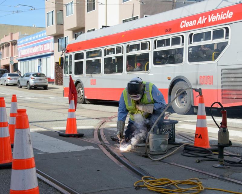 Crew member kneeling on the ground by the rail tracks. They are using a welder with several cones around them.