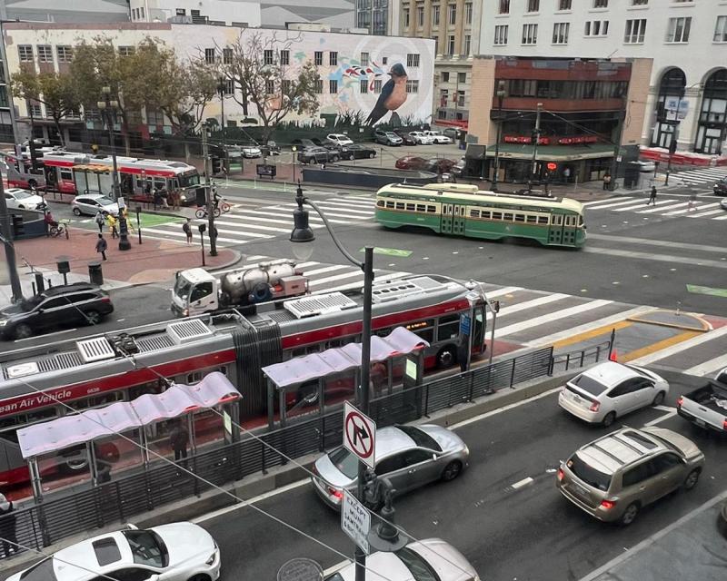 Image of a busy intersection corner possibly during commute showing multiple cars and buses, pedestrians and a trolley 