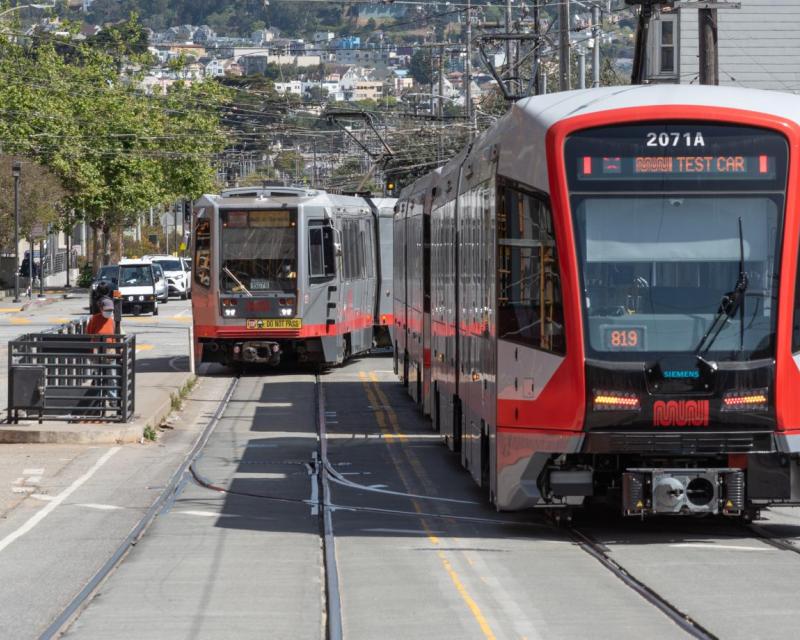 Photo of an older Breda model Muni Metro train on the street in the background, and a new LRV4 model Muni Metro train in the for