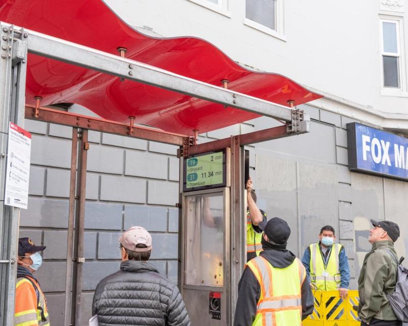  A picture containing contractors speaking with a Muni Rider as customer information displays are being installed in a transit s