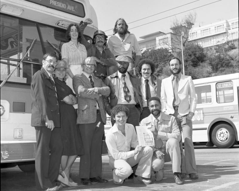 A dozen people dressed in business casual posting for a group photo in front of a bus in what seems to be a bus yard.  They are 