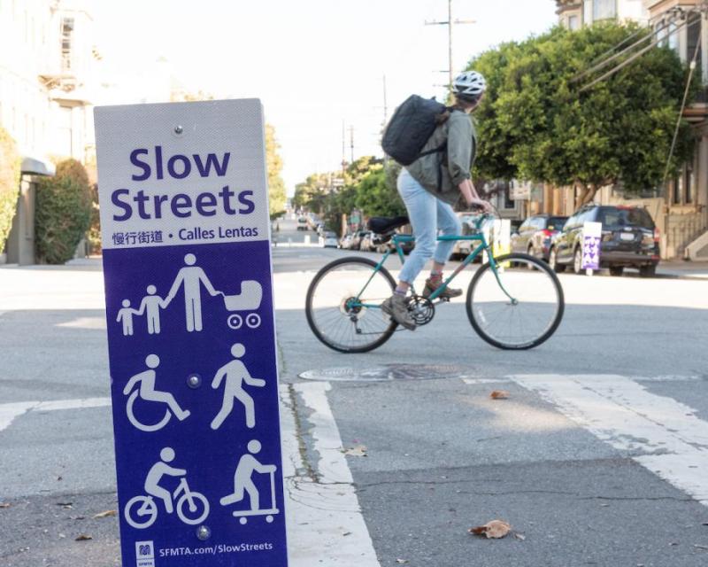 Close up image of a street sign with the words "Slow Streets" and multiple white figurine wheel users below. 