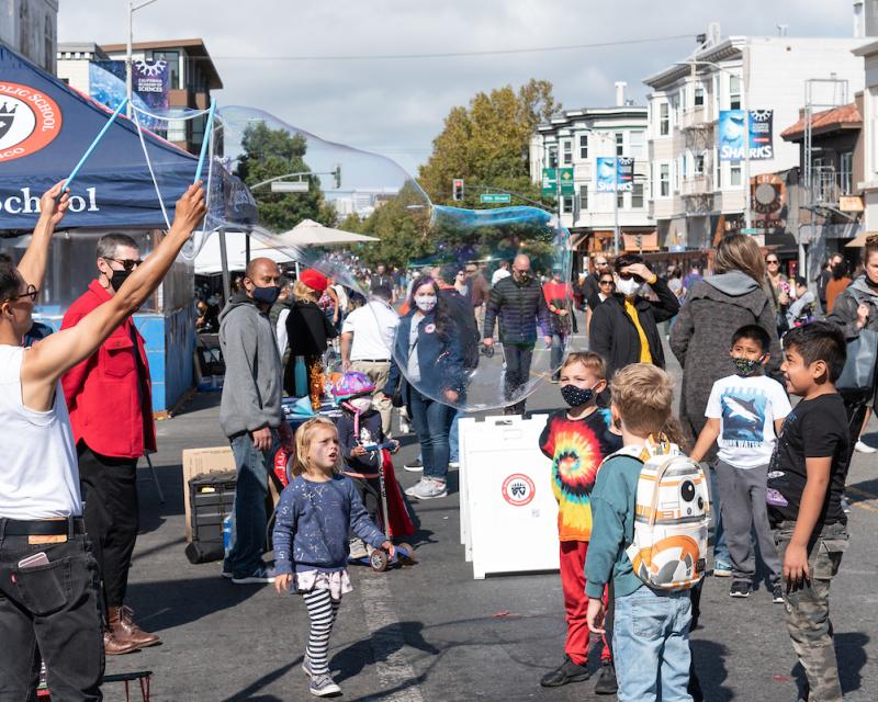 Children and adults seen on a street as a festive gathering, There are buildings in the background.