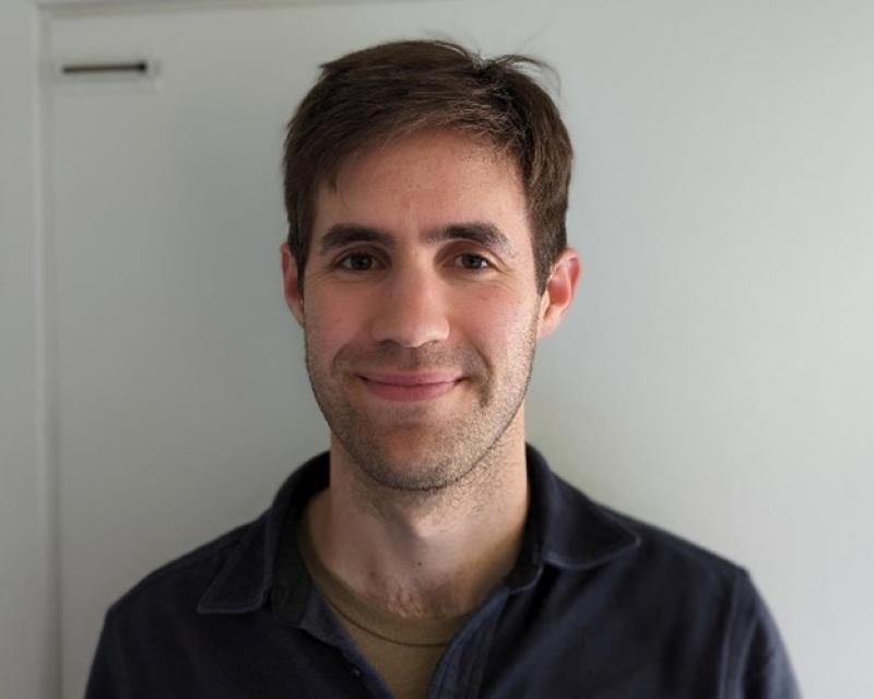 Man in a polo shirt smiling, standing in front of a white background wall.