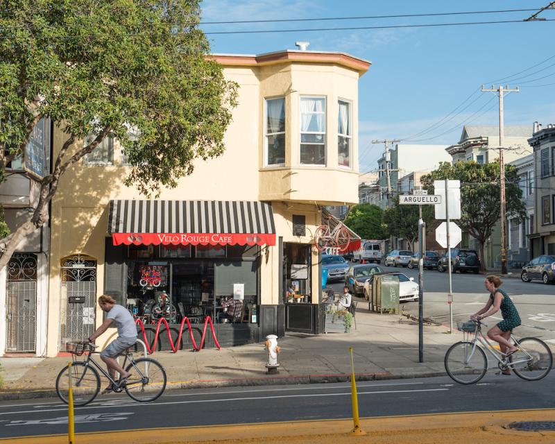 Two people on bicycles on the street ride in front of a restaurant with a person sitting outside.