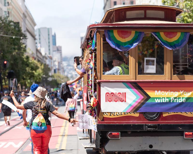 People march down the street at a parade around a historic cable car with rainbow flags.
