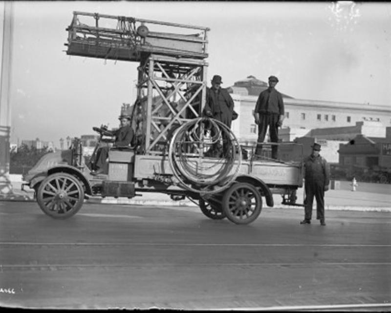 A black and white image of an old truck with a man in the driver seat, two in the in the truck bed, and one standing behind