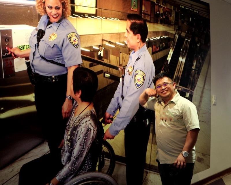 A man smiles in front of a picture of a three people with one of them in a wheelchair in front of an elevator.