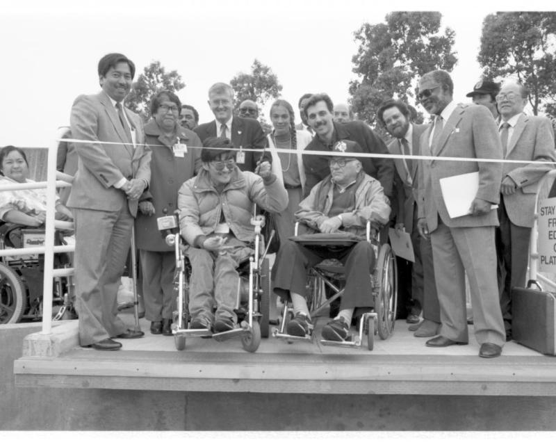 Black and white photo of people at a ribbon cutting ceremony. Two men in wheelchairs are in front to cut the ribbon.