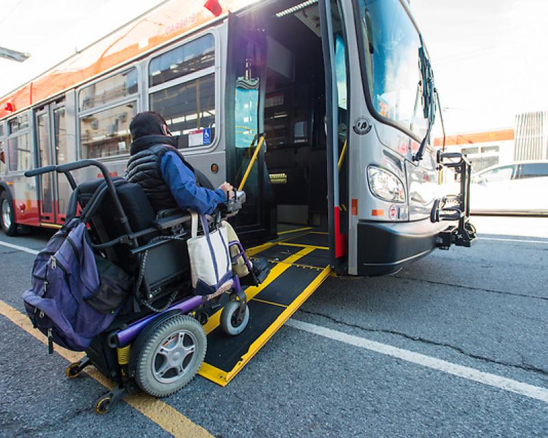A person in a blue long sleeved shirt and black vest using a motorized wheelchair and using a ramp to board a Muni bus.
