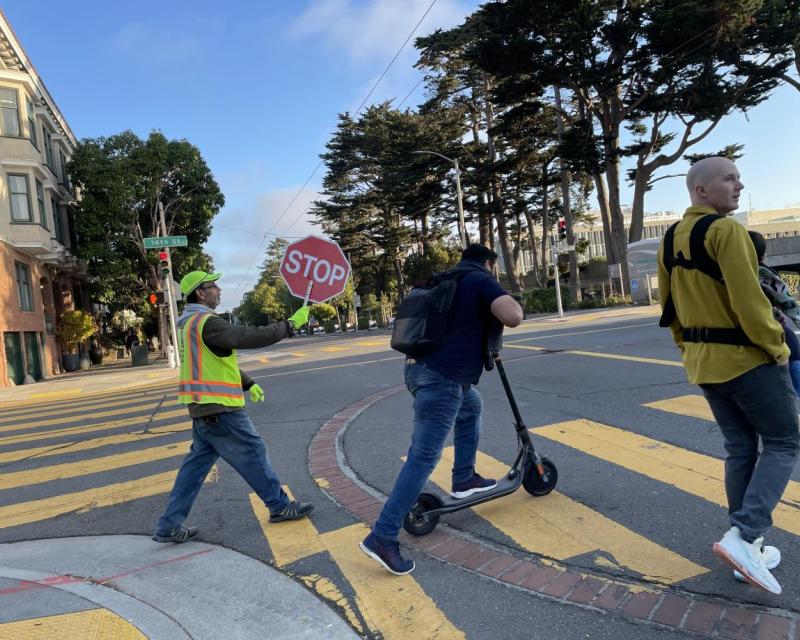 Three people in an intersection. One holding a stop sign, one on a scooter and one with a baby. 